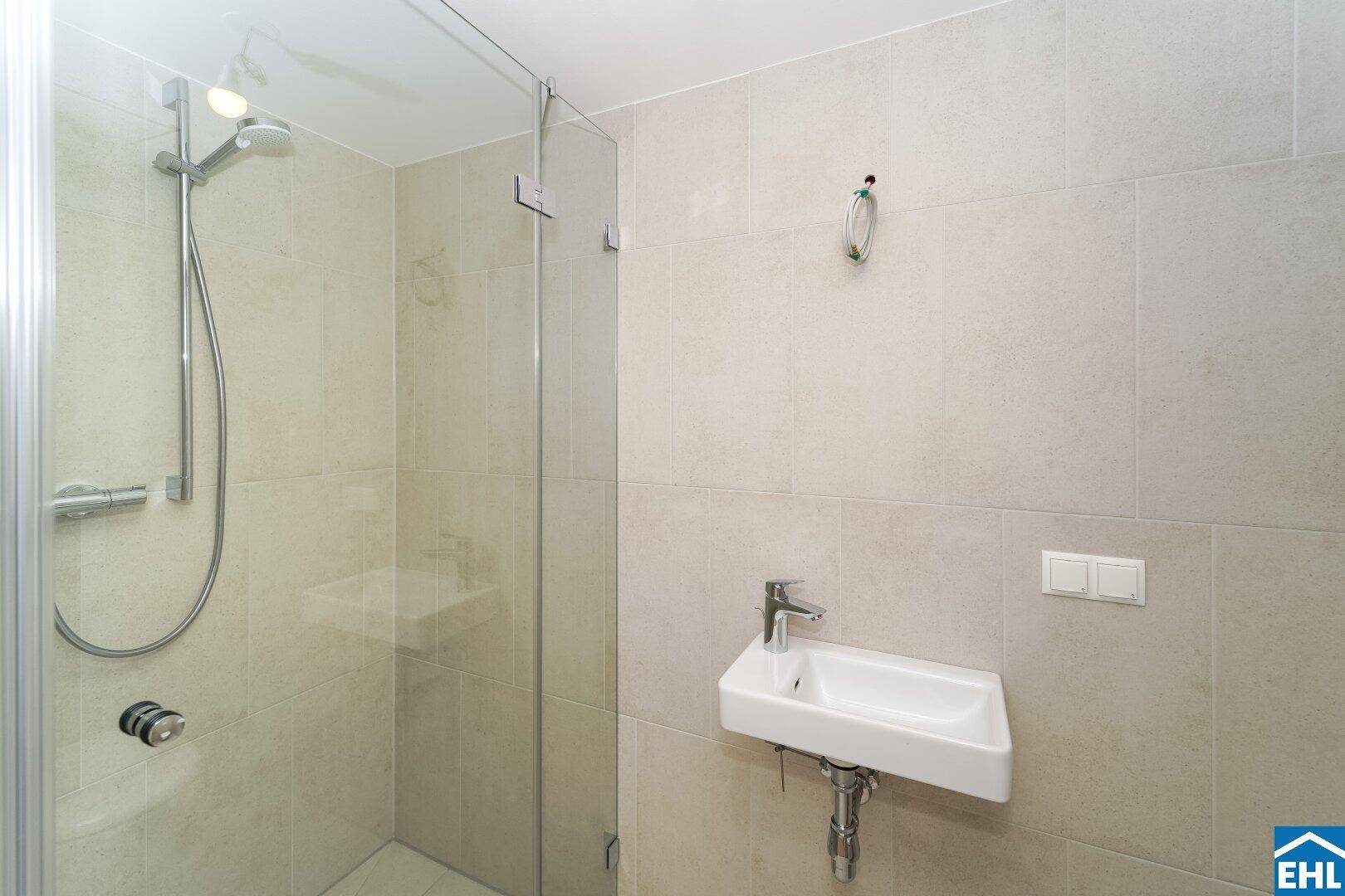Modernes Badezimmer mit Duschkabine, hellen Fliesen und kleinem Waschbecken.