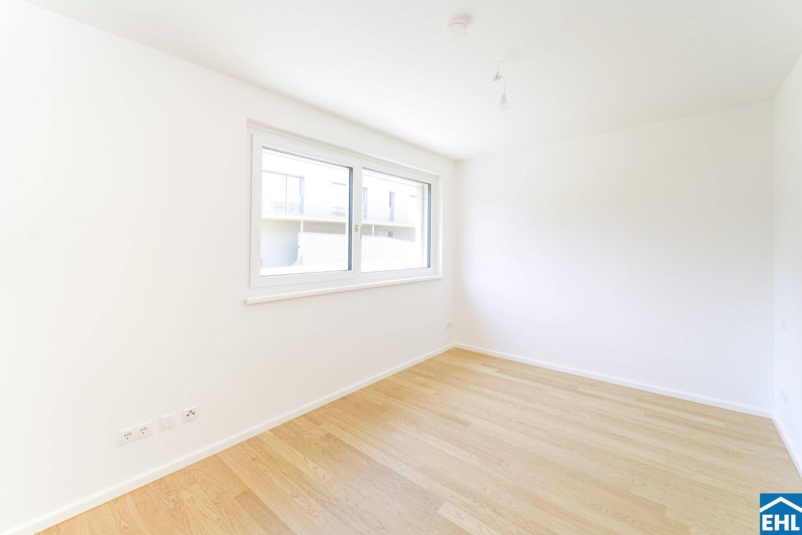 Helles, leeres Zimmer mit Parkettboden und einem großen Fenster.