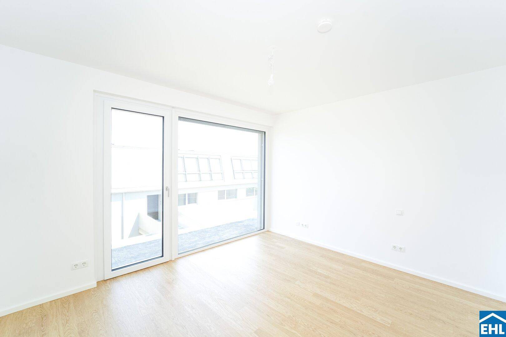 Heller Raum mit Parkettboden und großem Fenster mit Blick auf die umliegenden Gebäude.