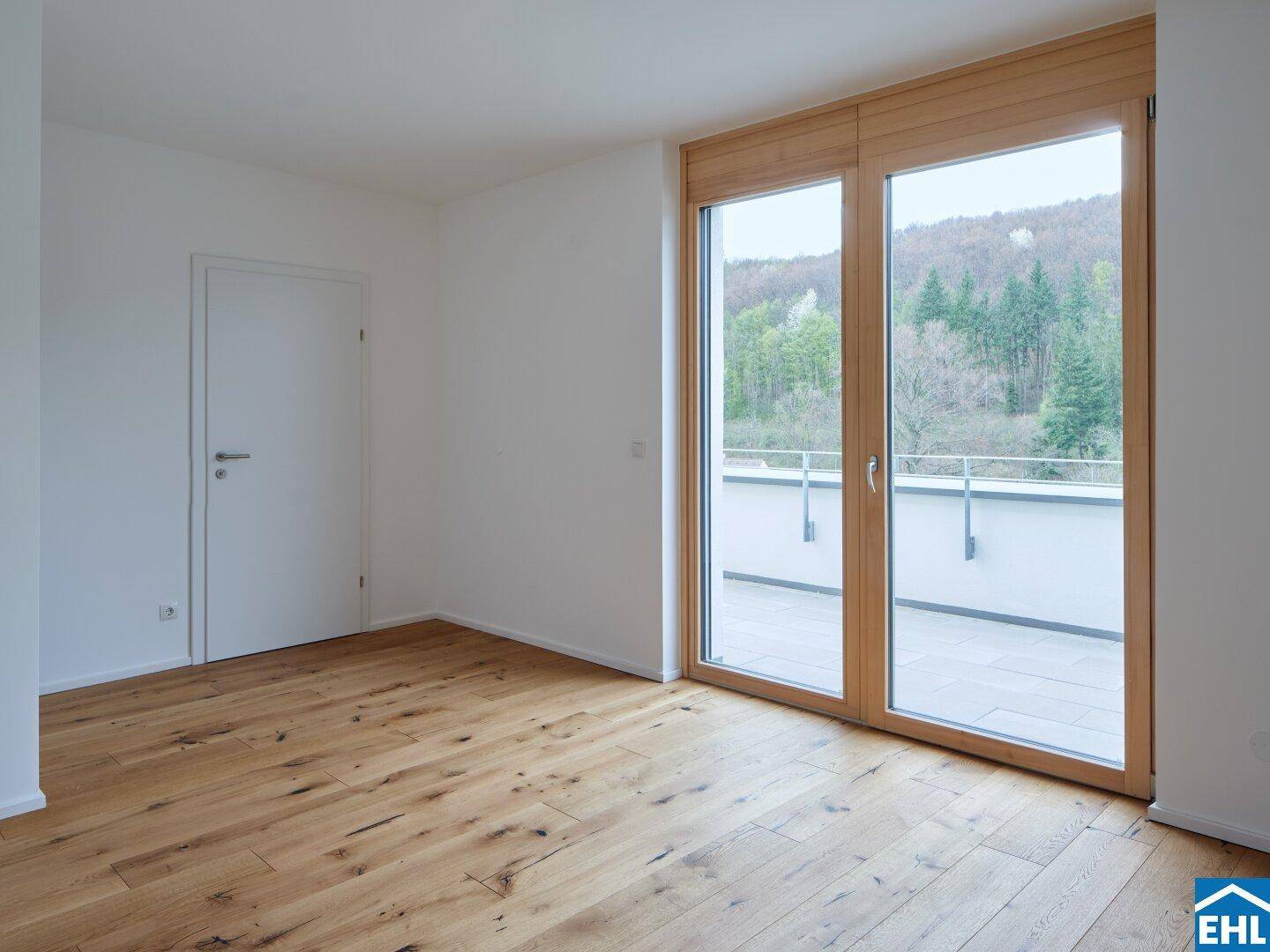 Leeres Zimmer mit Holzboden und großen Fenstern, die auf einen Balkon oder eine Terrasse mit Grünblick führen.