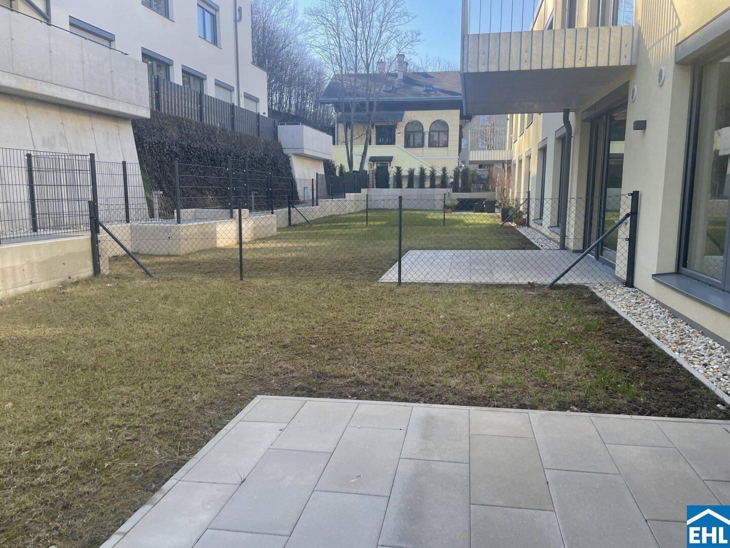 Gepflegter Gartenbereich mit Rasenfläche und gepflasterter Terrasse, umgeben von Zäunen und Gebäuden.