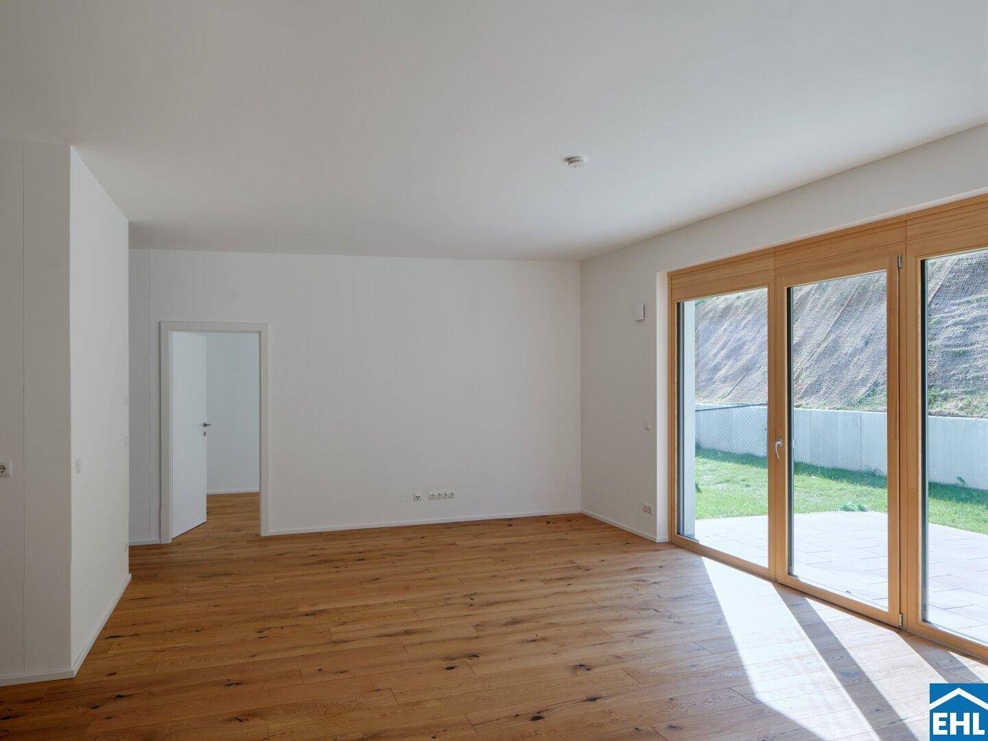 Helles, leeres Wohnzimmer mit Holzboden und großen Fenstern, die Zugang zu einem Außenbereich bieten.