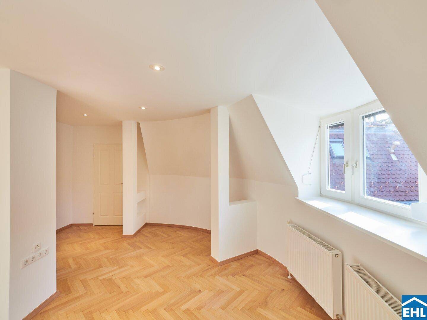 Heller Dachgeschossraum mit schrägen Wänden, Fischgrätparkett und einem Dachfenster für zusätzliches Licht.