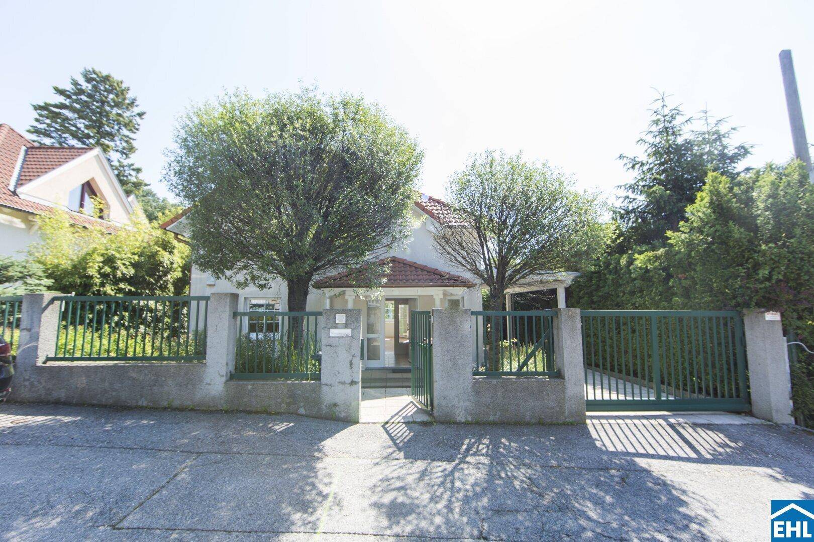 Ansicht des Hauses von der Straße mit grünem Zaun und Einfahrtstor, umgeben von Bäumen.
