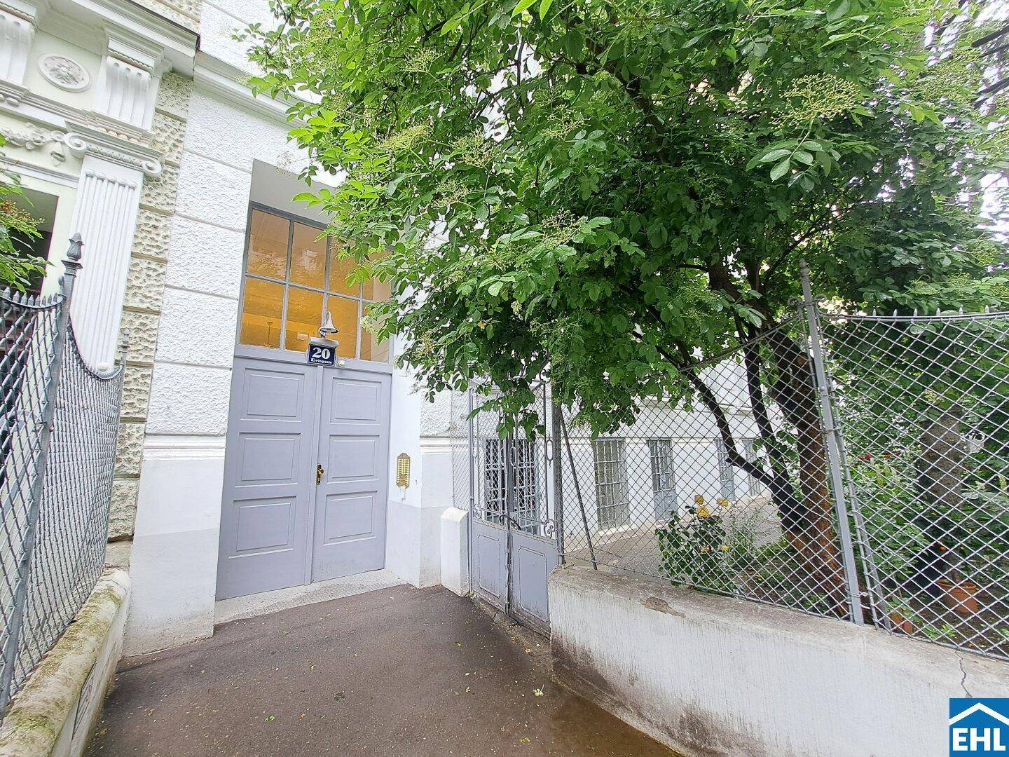 Eingangsbereich eines Gebäudes mit grauer Doppeltür, Hausnummer 20 und einem Baum davor.