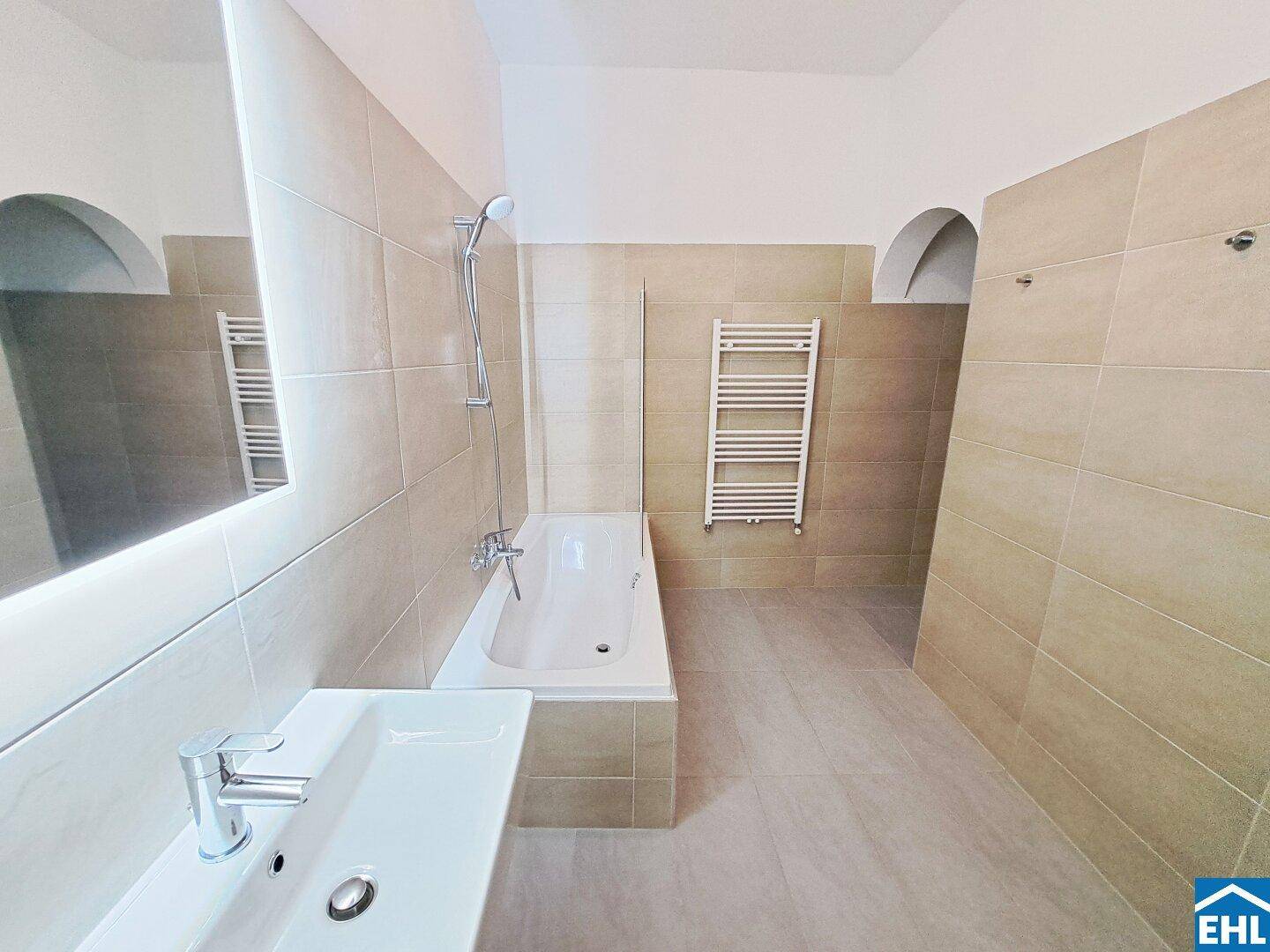 Geräumiges Badezimmer mit beigen Fliesen, Badewanne, Waschbecken und beheiztem Handtuchhalter.
