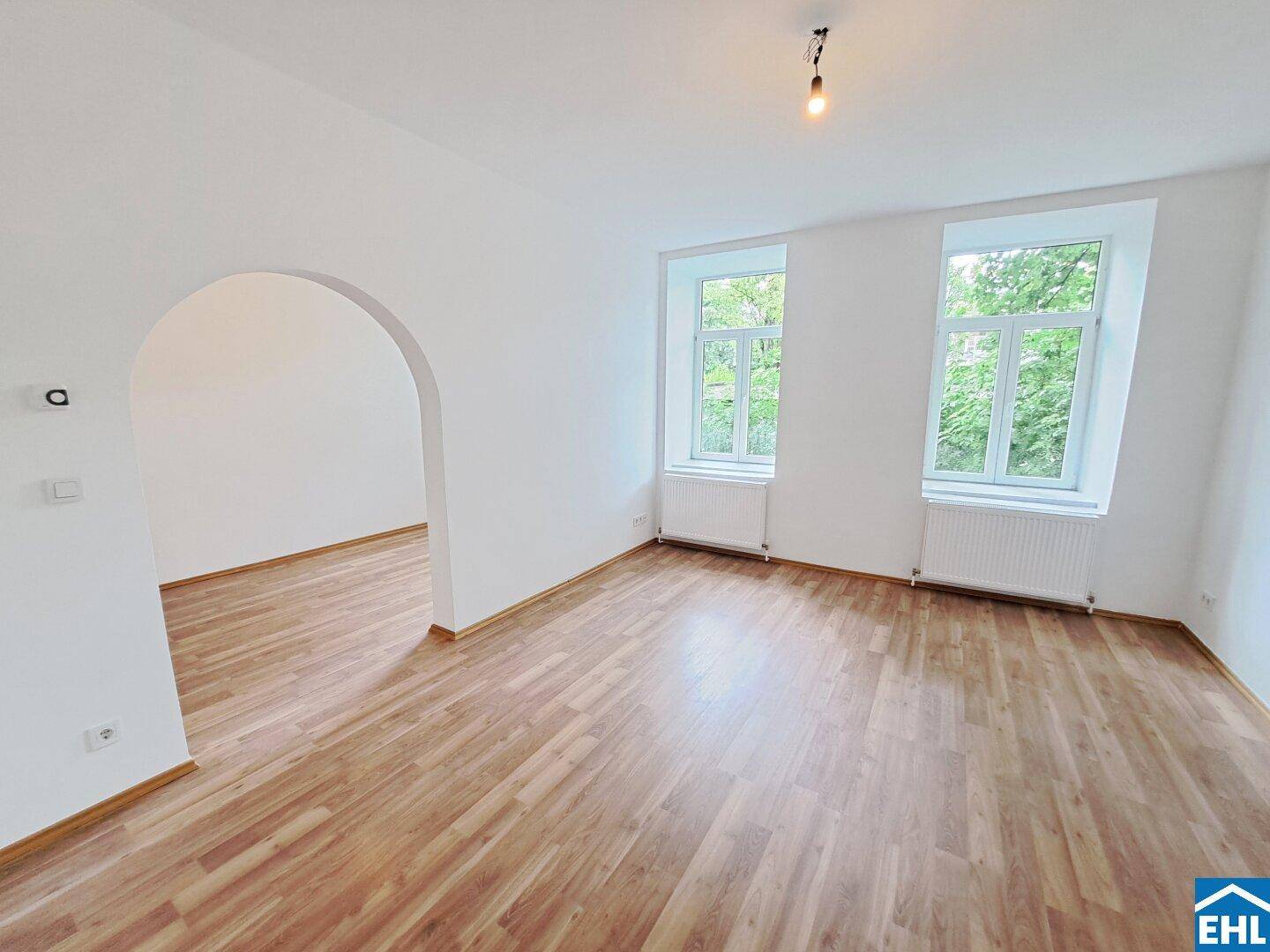 Helles Wohnzimmer mit Laminatboden, weißen Wänden und Blick ins Grüne durch große Fenster.
