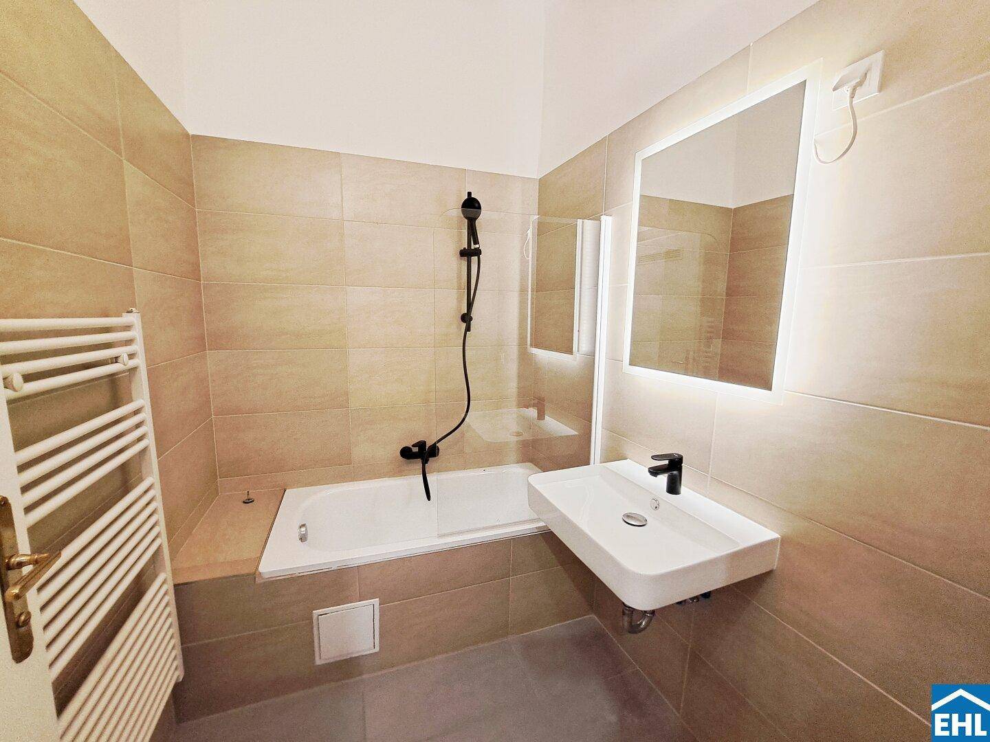 Modernes Badezimmer mit beigen Fliesen, Badewanne, beleuchtetem Spiegel und beheiztem Handtuchhalter.