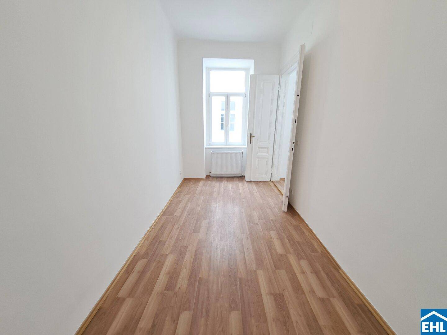 Leeres Zimmer mit Laminatboden und einem Fenster am Ende des Raumes, ideal als Schlafzimmer.