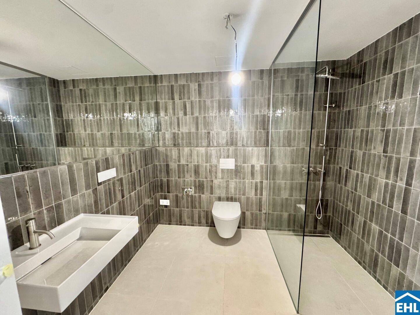 Modernes Badezimmer mit grauen Fliesen, wandhängendem WC und einer geräumigen begehbaren Dusche.