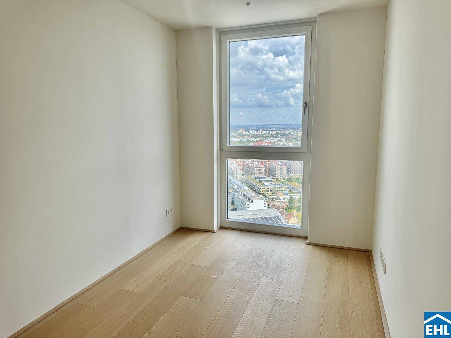 Schlichter Raum mit Holzboden und einem hohen Fenster, das viel Tageslicht und Stadtblick hereinlässt.