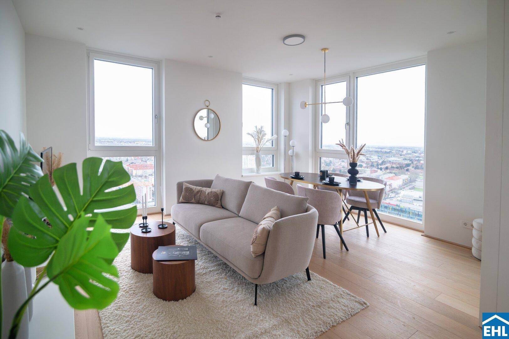 Heller Wohn- und Essbereich mit beigem Sofa, Holzboden und großen Fenstern, die viel Licht hereinlassen.