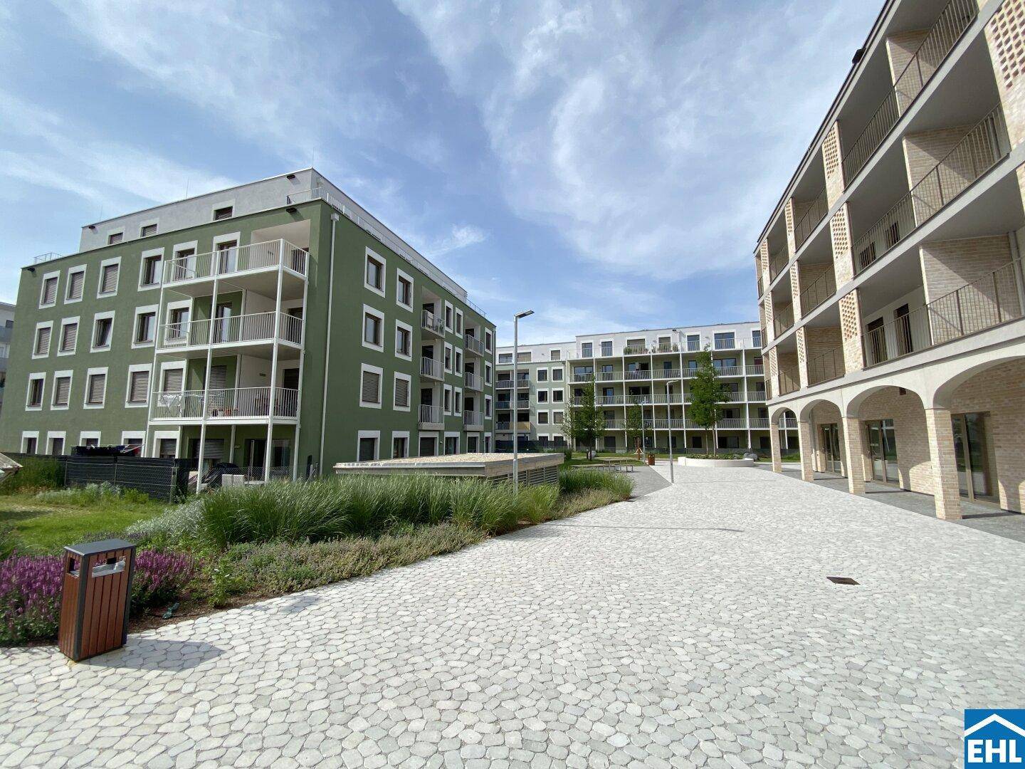 Weitläufiger Innenhof mit gepflasterten Wegen, Grünflächen und modernen Wohngebäuden unter blauem Himmel.