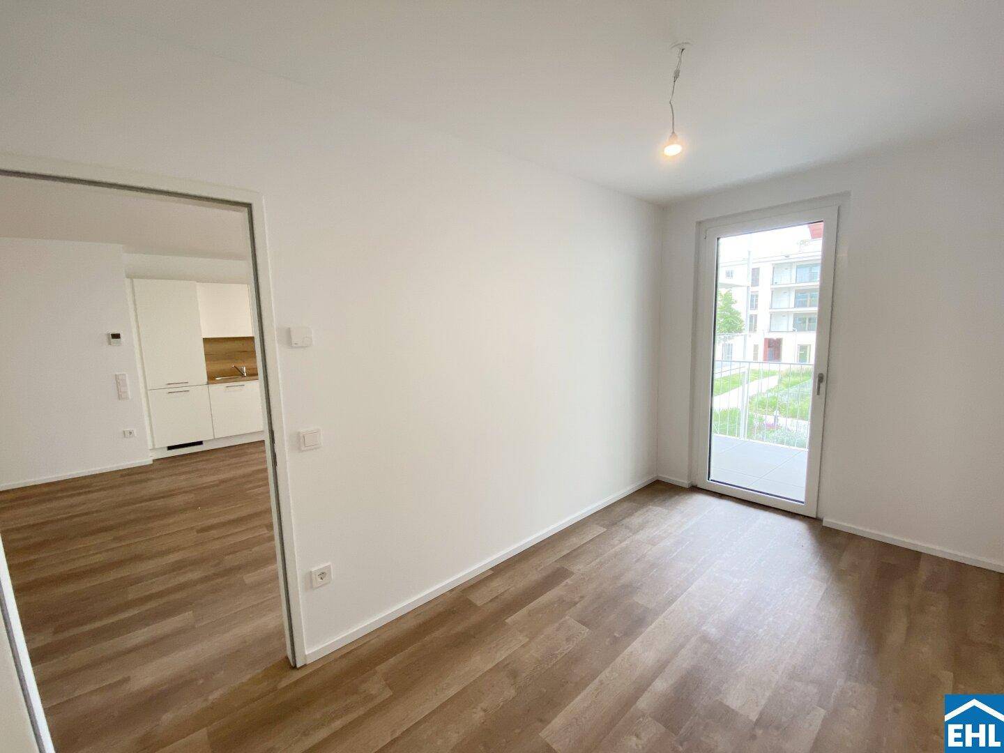 Heller Raum mit Laminatboden und Zugang zu einem Balkon, Blick in die offene Küche.