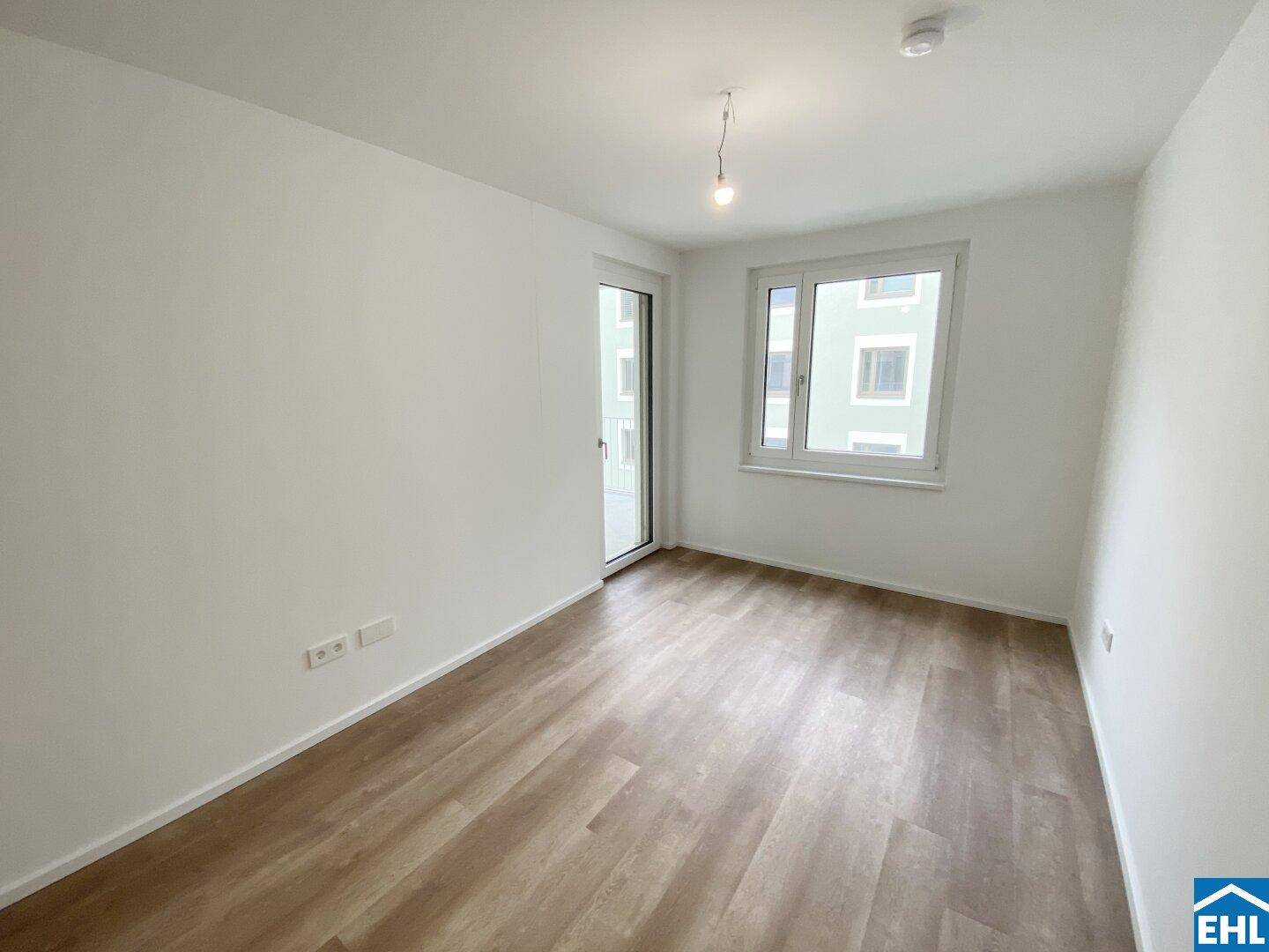 Leeres Schlafzimmer mit Laminatboden, großen Fenstern und Zugang zum Balkon.