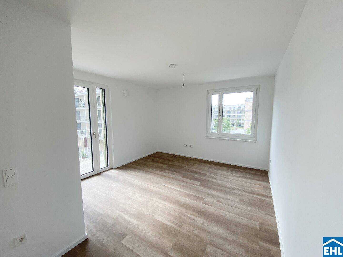 Heller, leerer Raum mit Laminatboden, großen Fenstern und Zugang zu einem Balkon.