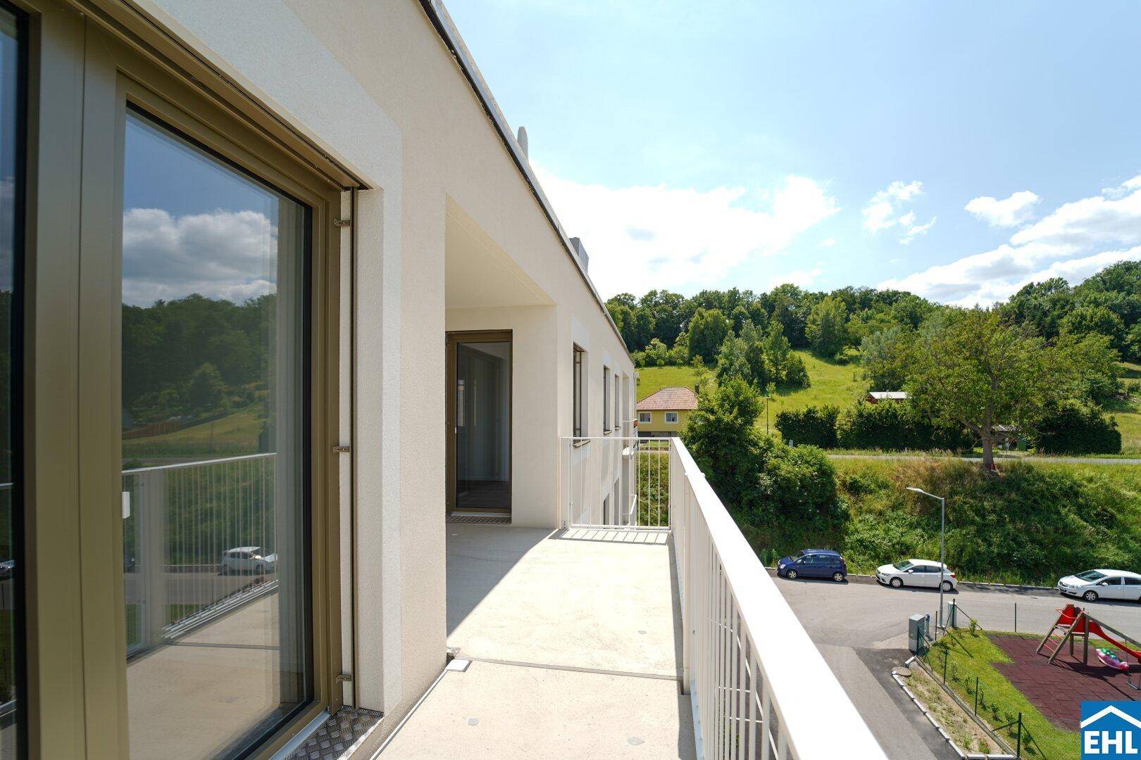 Geräumiger Balkon mit weitem Blick auf grüne Hügel, eine Straße und einen Kinderspielplatz.