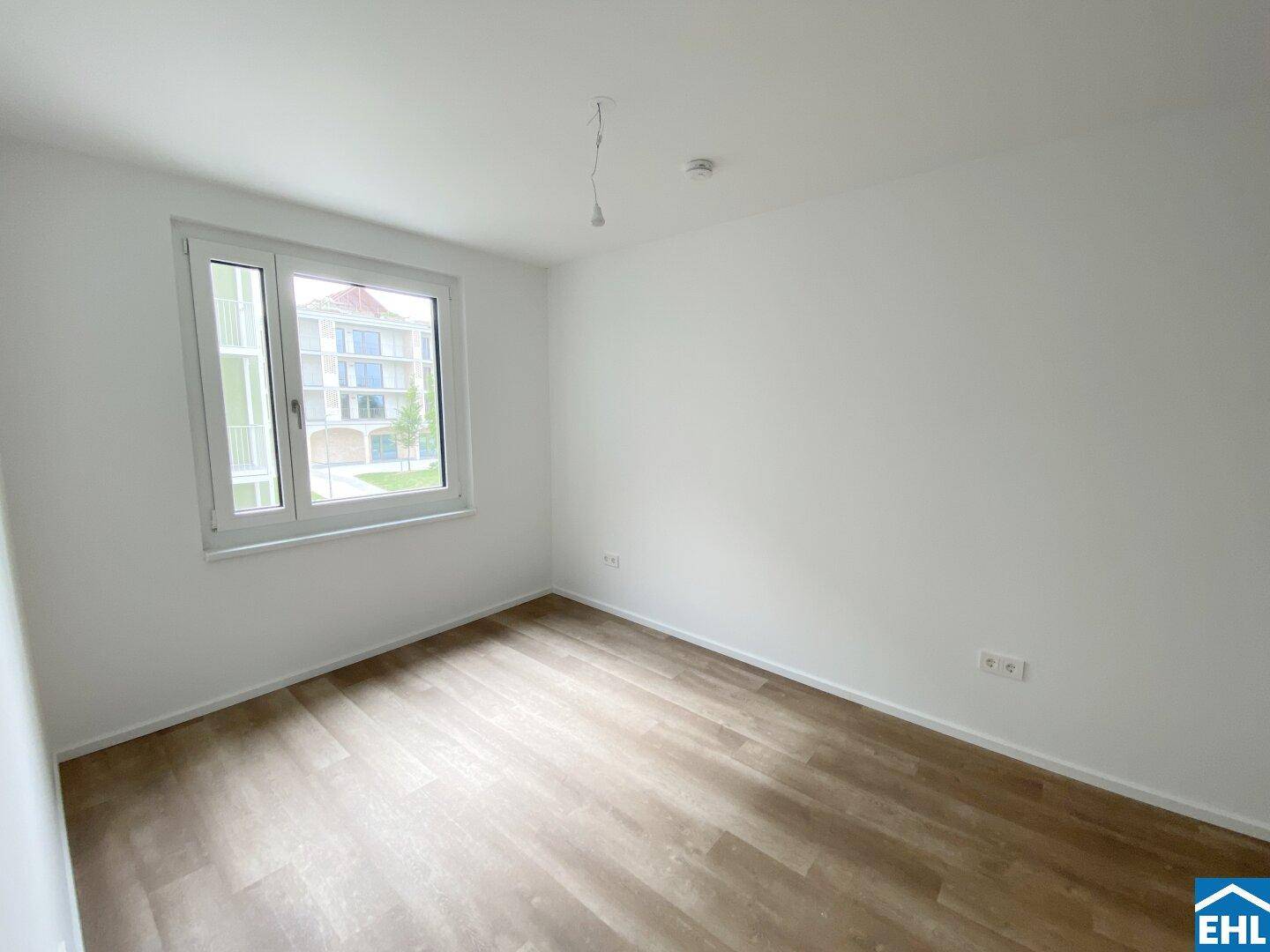 Ruhiges Schlafzimmer mit Laminatboden und einem Fenster, das den Blick auf ein benachbartes Gebäude freigibt.