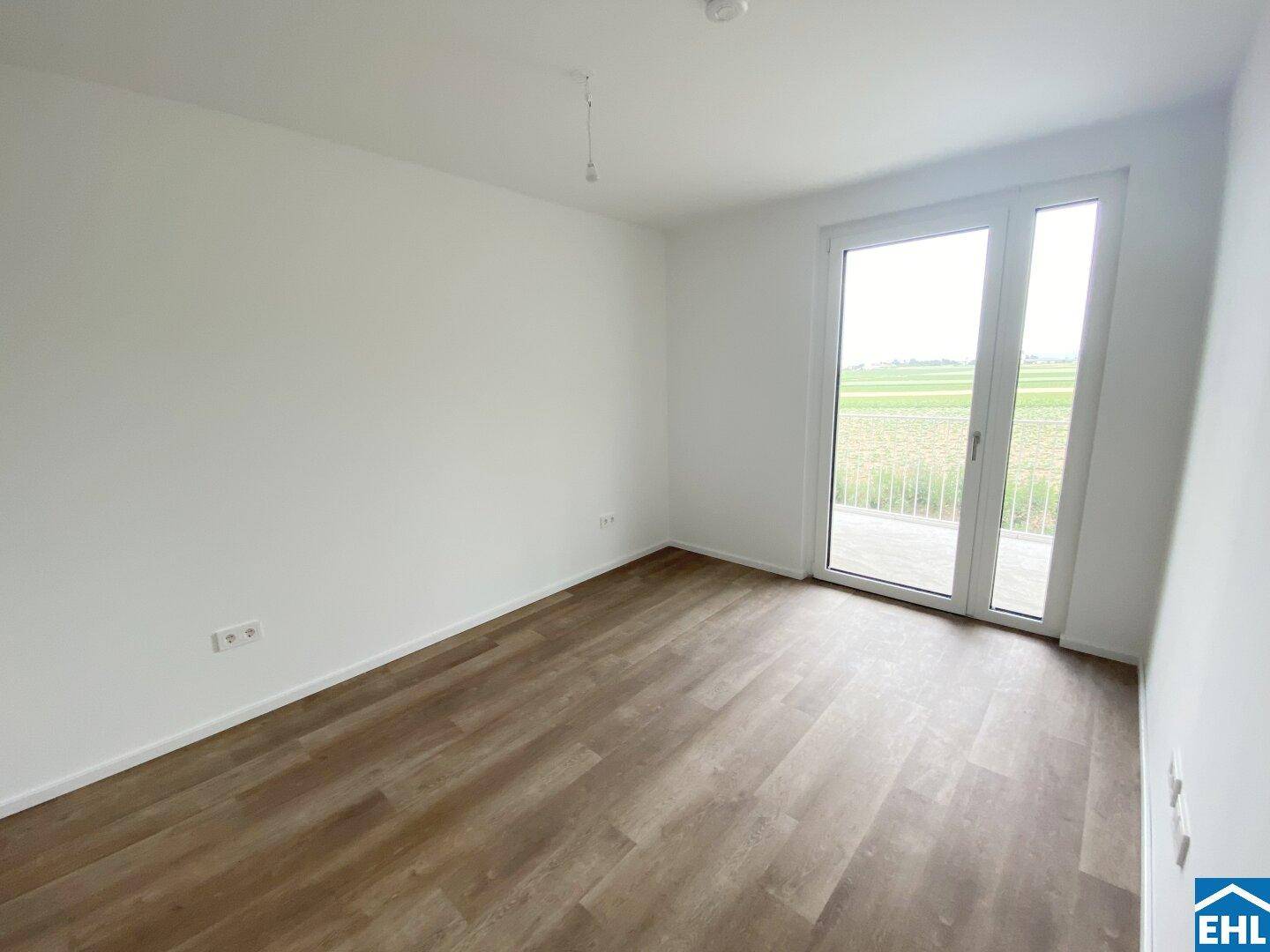 Helles Zimmer mit Laminatboden und Zugang zu einem Balkon mit weitem Blick ins Grüne.