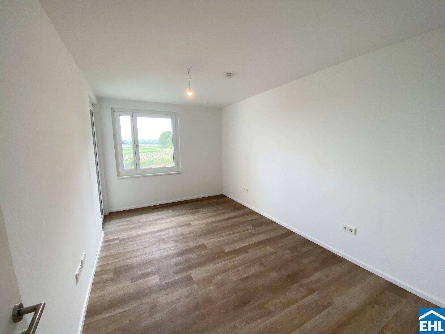 Helles, leeres Zimmer mit modernem Laminatboden und Blick ins Grüne.
