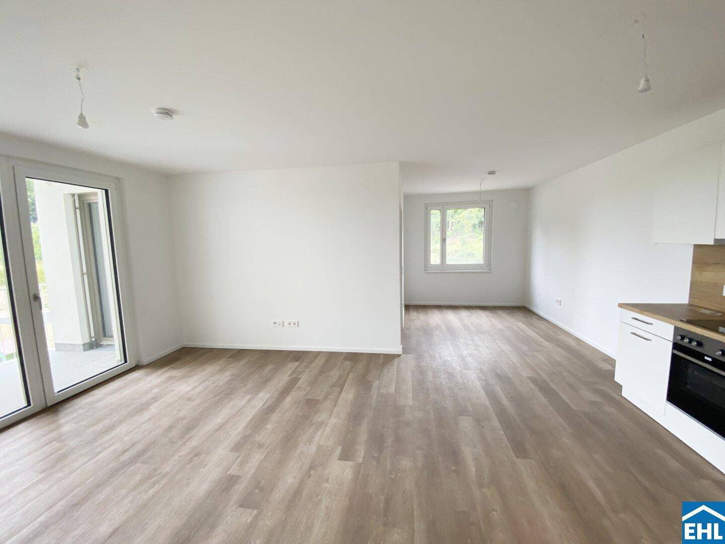 Offener Wohnbereich mit Laminatboden, Zugang zum Balkon und teilweiser Sicht auf die Küche.