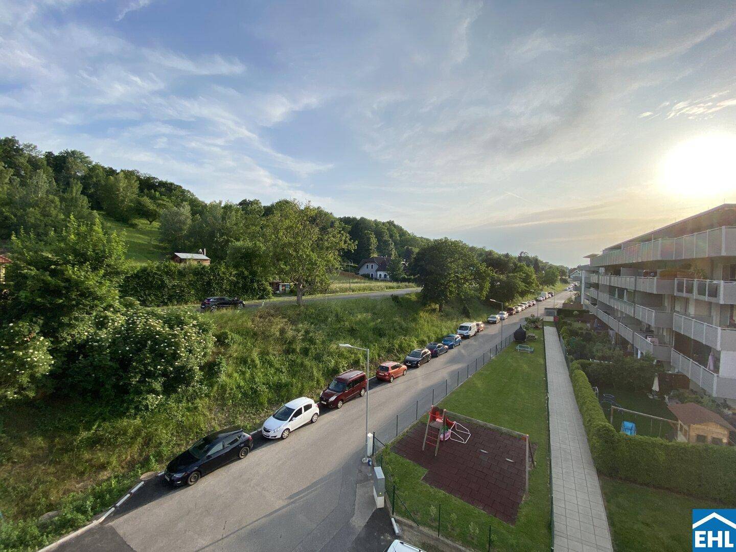 Weitläufiger Blick über eine Straße, grüne Hügel und die umliegende Wohnbebauung bei Sonnenuntergang.