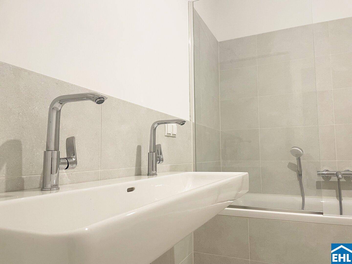 Modernes Badezimmer mit breitem Waschbecken, zwei Armaturen und Badewanne mit Duschfunktion.