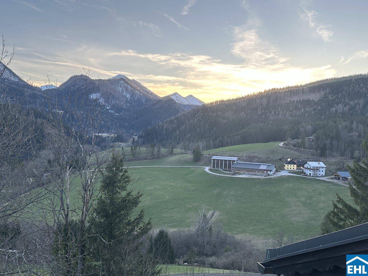 Malerischer Blick auf eine weite Landschaft mit Bergen, Tälern und vereinzelten Gebäuden bei Sonnenuntergang.