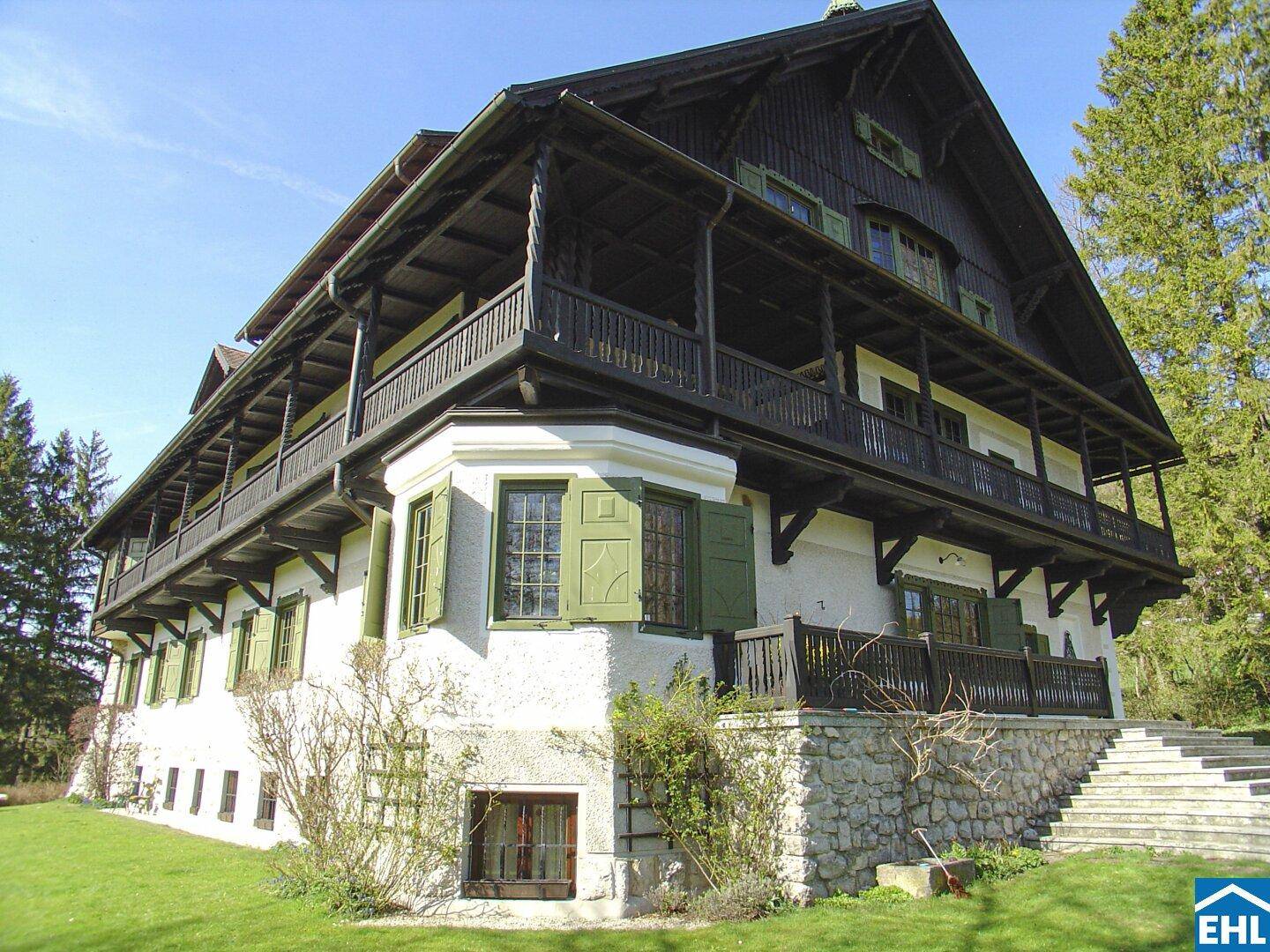 Traditionelles Landhaus mit dunkler Holzverkleidung und grünen Fensterläden, umgeben von Natur.