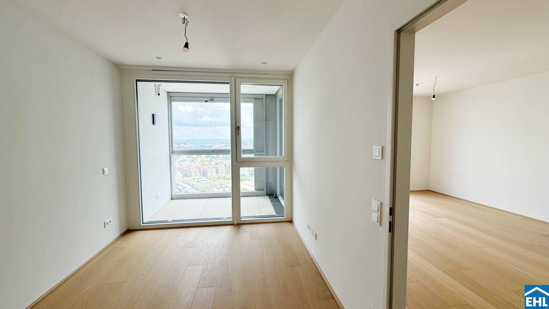 Heller Raum mit Holzboden und Zugang zu einem Balkon, der einen weiten Blick über die Stadt ermöglicht.