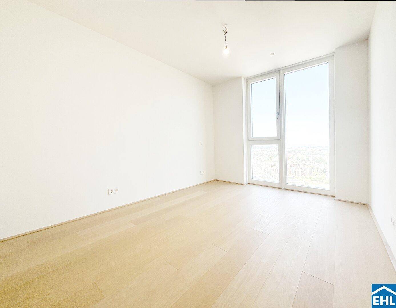 Leeres Schlafzimmer mit hellem Holzboden und großem Fenster, das viel Tageslicht hereinlässt.