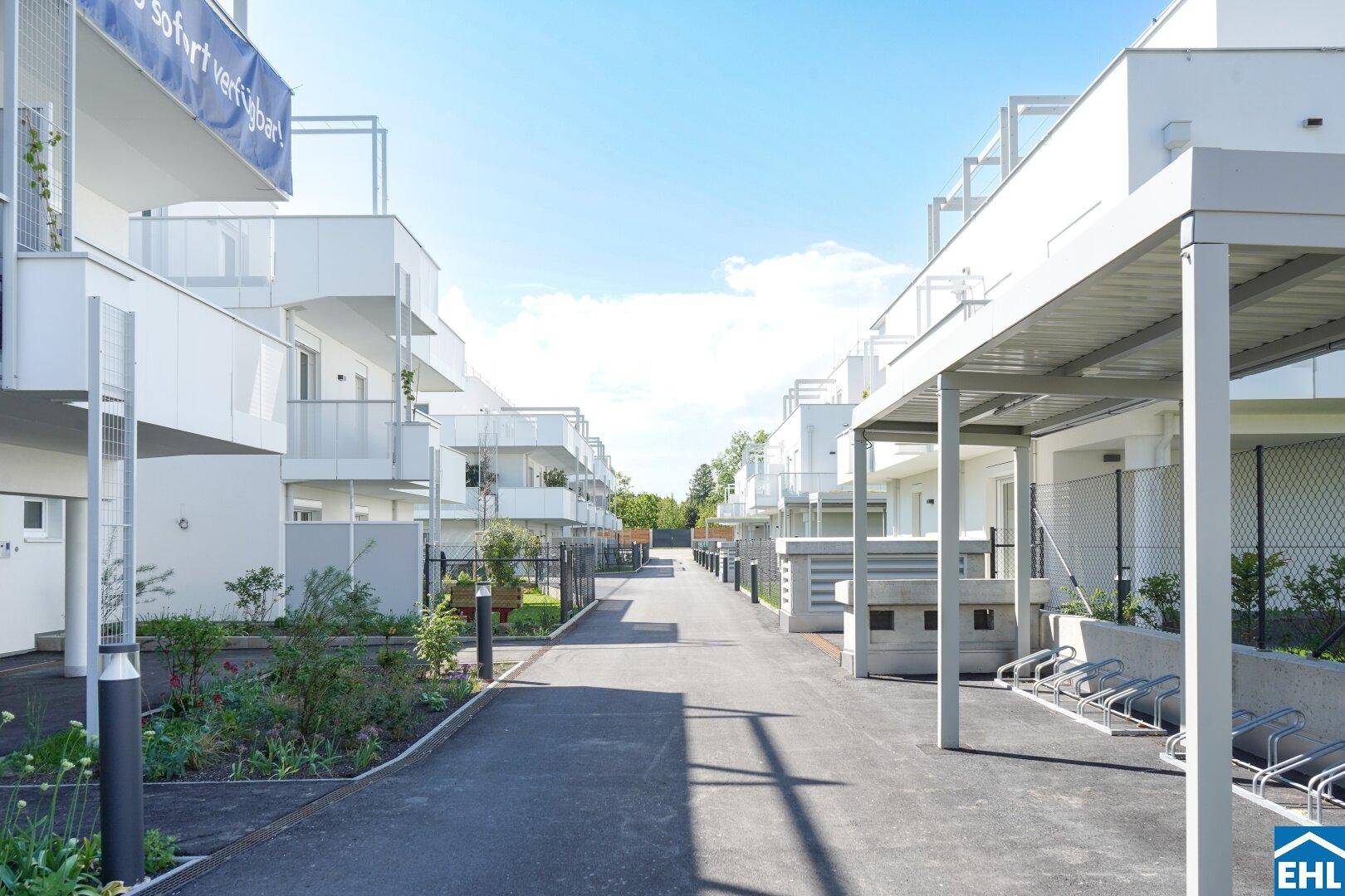 Eine Reihe moderner, weißer Wohngebäude entlang eines asphaltierten Weges mit Grünflächen und Carports.