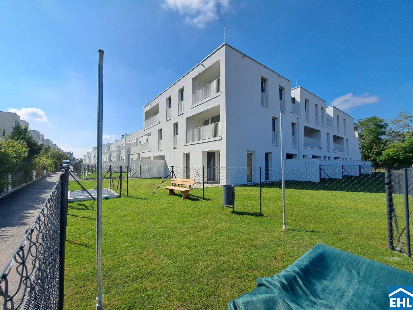 Eckgebäude des Neubaus mit angrenzender Grünfläche und Sitzbank bei Sonnenschein und klarem Himmel.