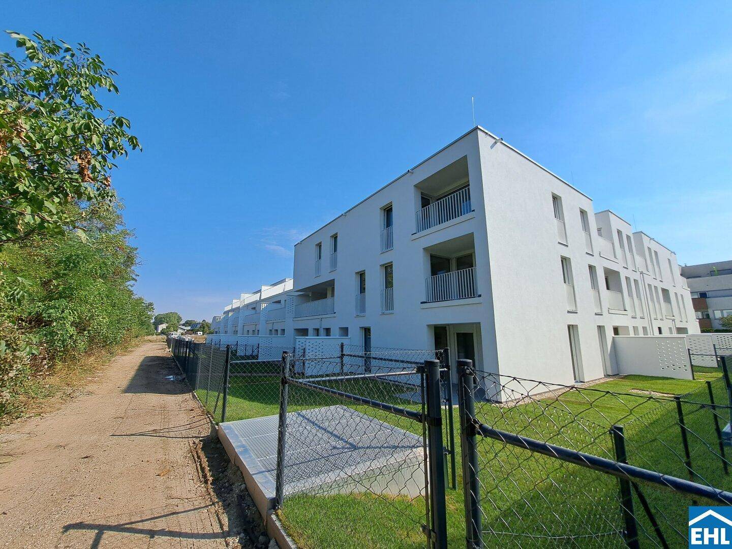 Moderne weiße Wohngebäude mit Balkonen und kleinen Gärten unter blauem Himmel.