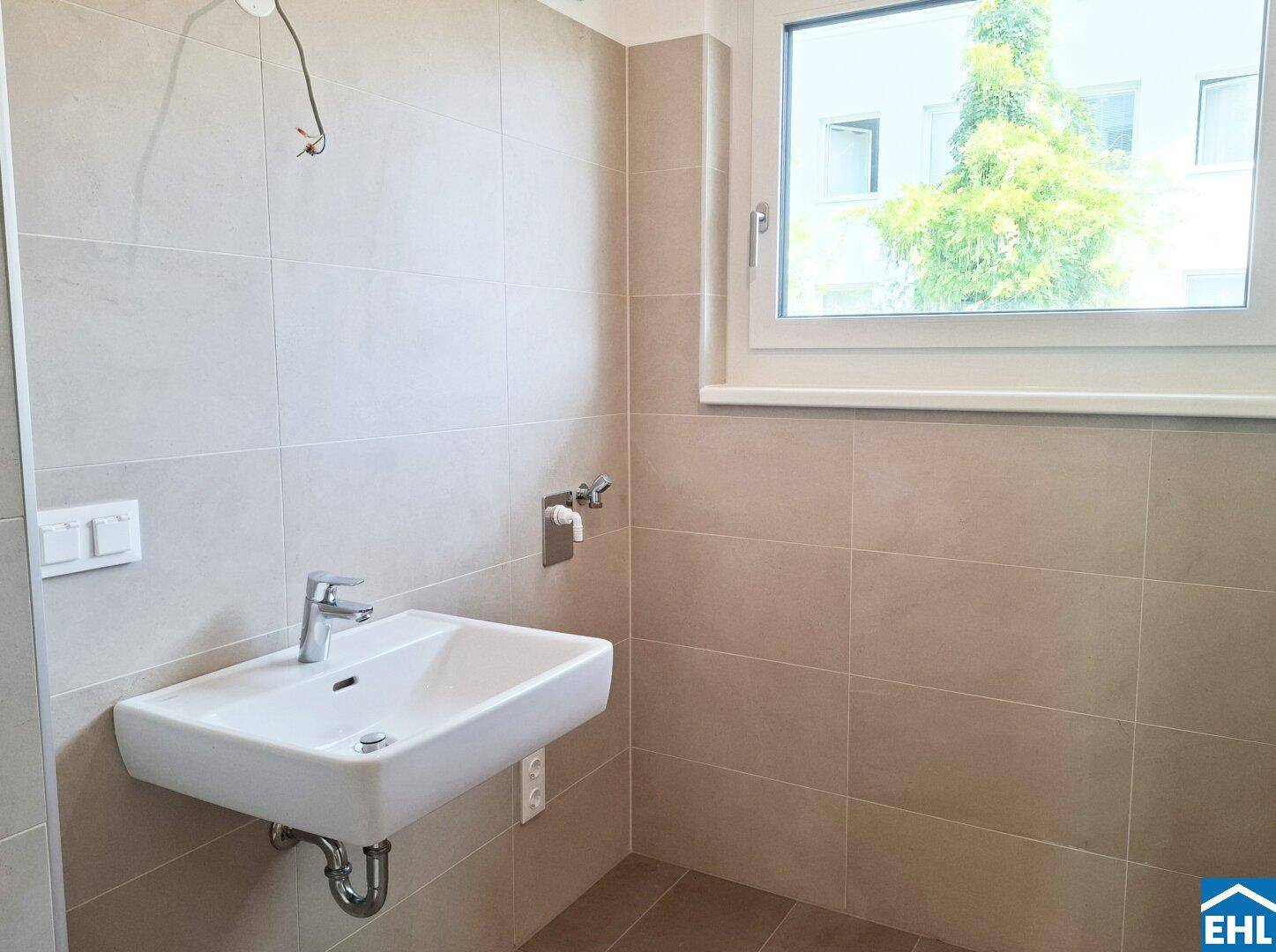 Helles Badezimmer mit Einzelwaschbecken, Fenster und beigefarbenen Fliesen, unmöbliert.