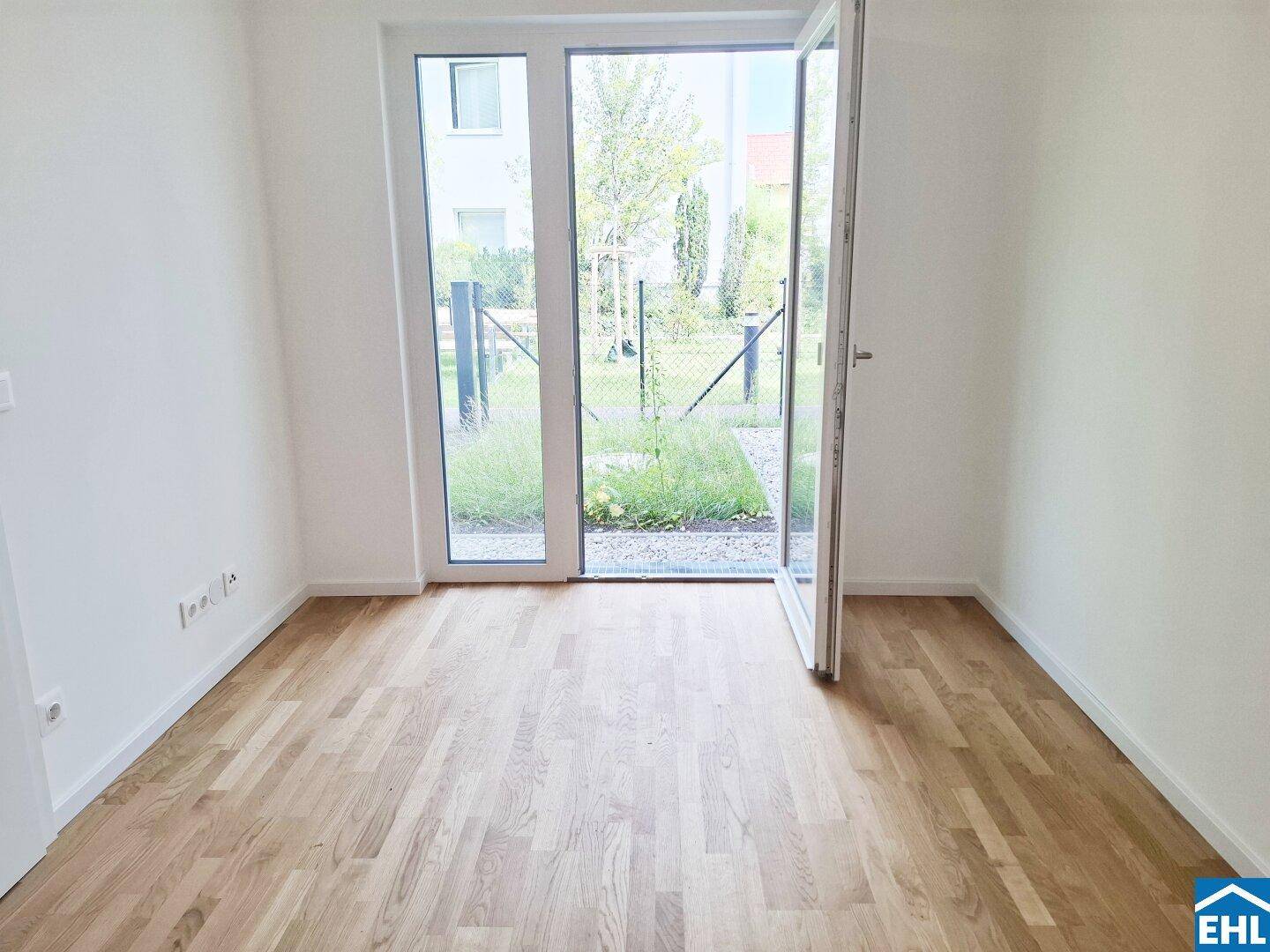 Leerer Raum mit Holzboden und großen Fenstertüren, die zum grünen Garten führen.
