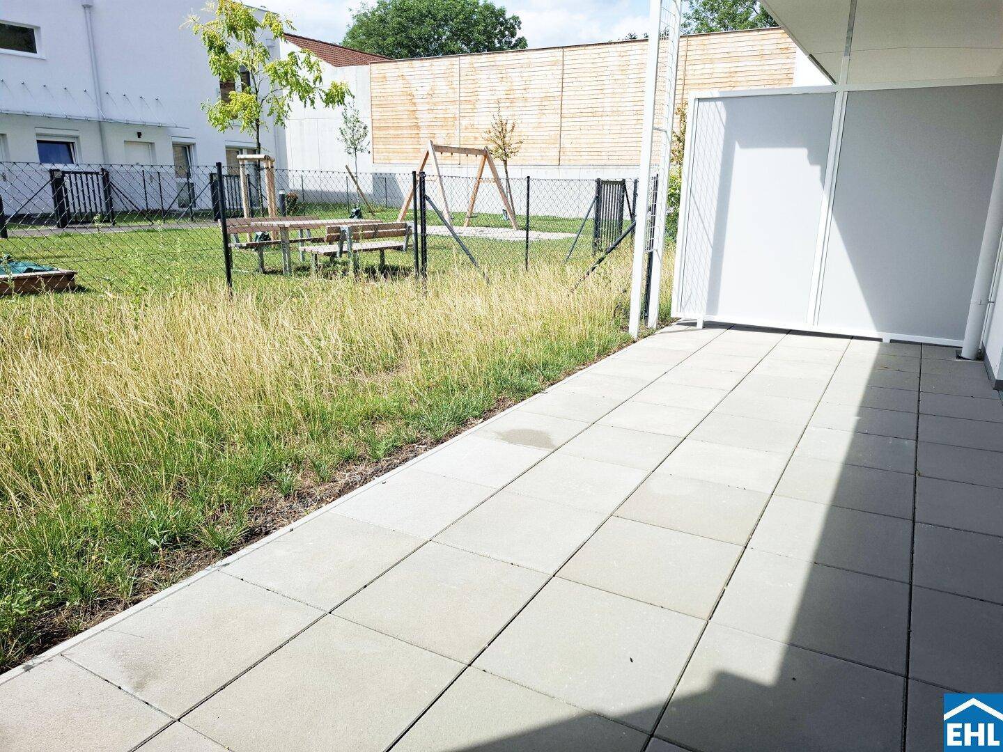 Gepflasterte Terrasse neben einer Rasenfläche, umgeben von einem Zaun.