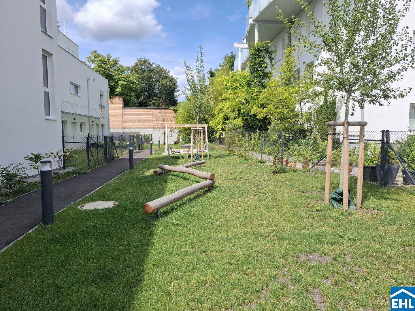 Grüne Gemeinschaftsfläche mit Holzspielgeräten und jungen Bäumen für Kinder in der Wohnanlage.