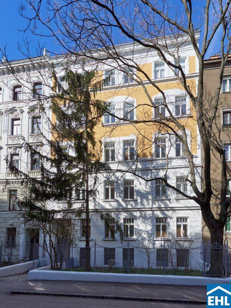 Historisches Gebäude mit weißer und gelber Fassade, umgeben von Bäumen und einem Zaun.