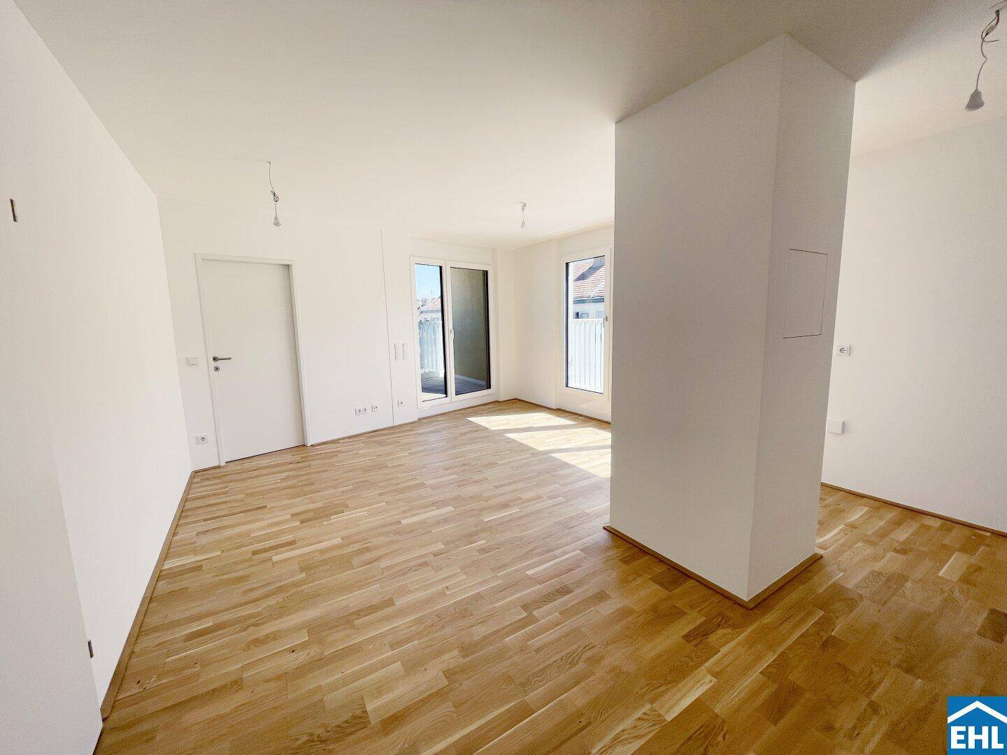 Helles, leeres Zimmer mit Holzboden und großen Fenstern, die viel Tageslicht hereinlassen.