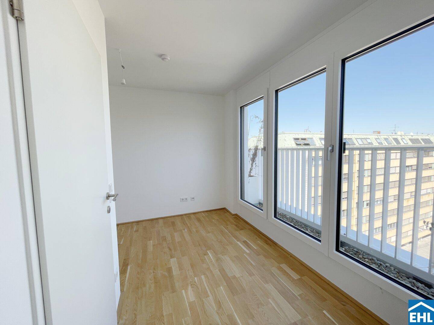 Helles Zimmer mit Holzboden und großen Fenstern, die einen Blick auf die Stadt und einen Balkon bieten.