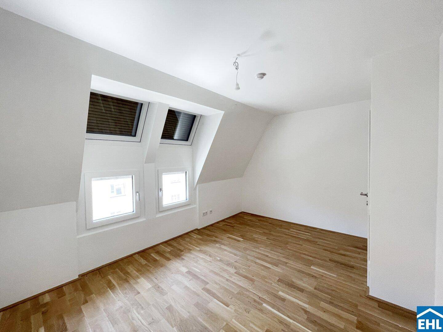 Dachzimmer mit Holzboden, weißen Wänden und schrägen Fenstern, die mit Jalousien ausgestattet sind.