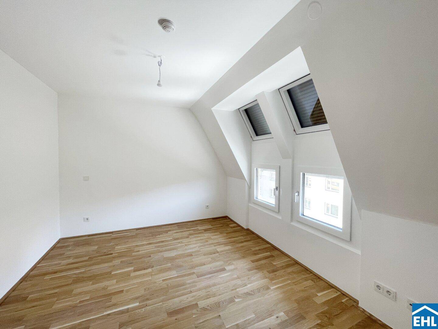 Geräumiges Dachzimmer mit Holzboden und schrägen Wänden, ausgestattet mit Dachfenstern und Jalousien.