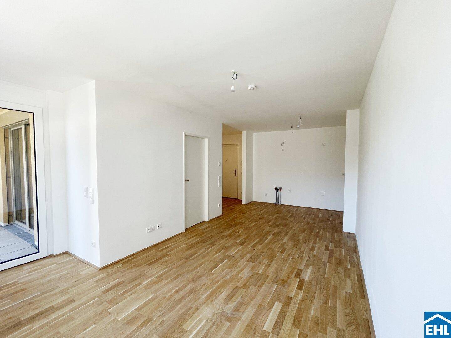 Lichtdurchfluteter Raum mit Parkettboden, weißen Wänden und Anschlussmöglichkeiten für eine Küche.