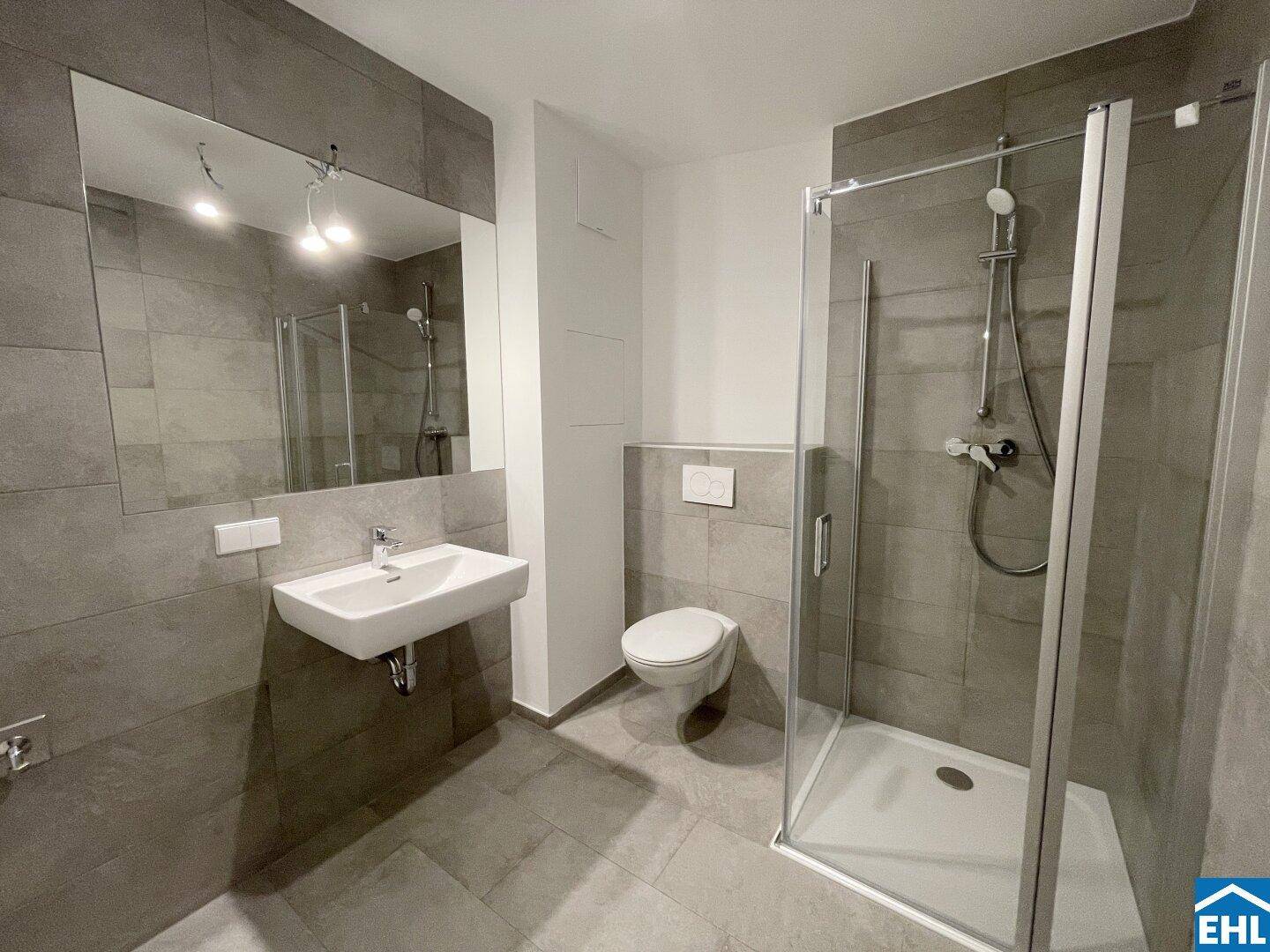 Modernes Badezimmer mit grauen Fliesen, wandhängendem WC und einer geräumigen begehbaren Dusche.