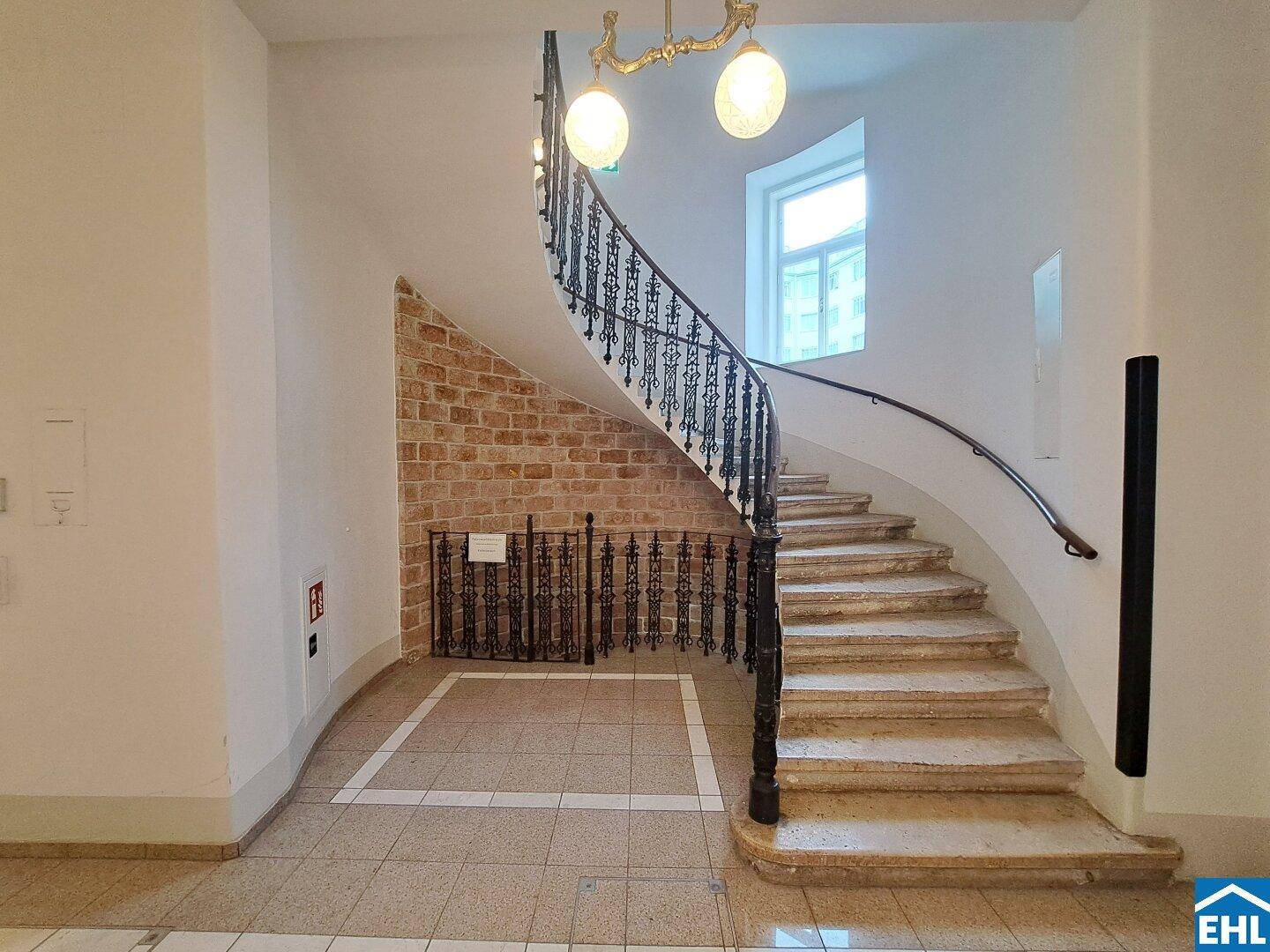 Elegantes Treppenhaus mit geschwungener Steintreppe, schmiedeeisernem Geländer und einer dekorativen Ziegelwand.