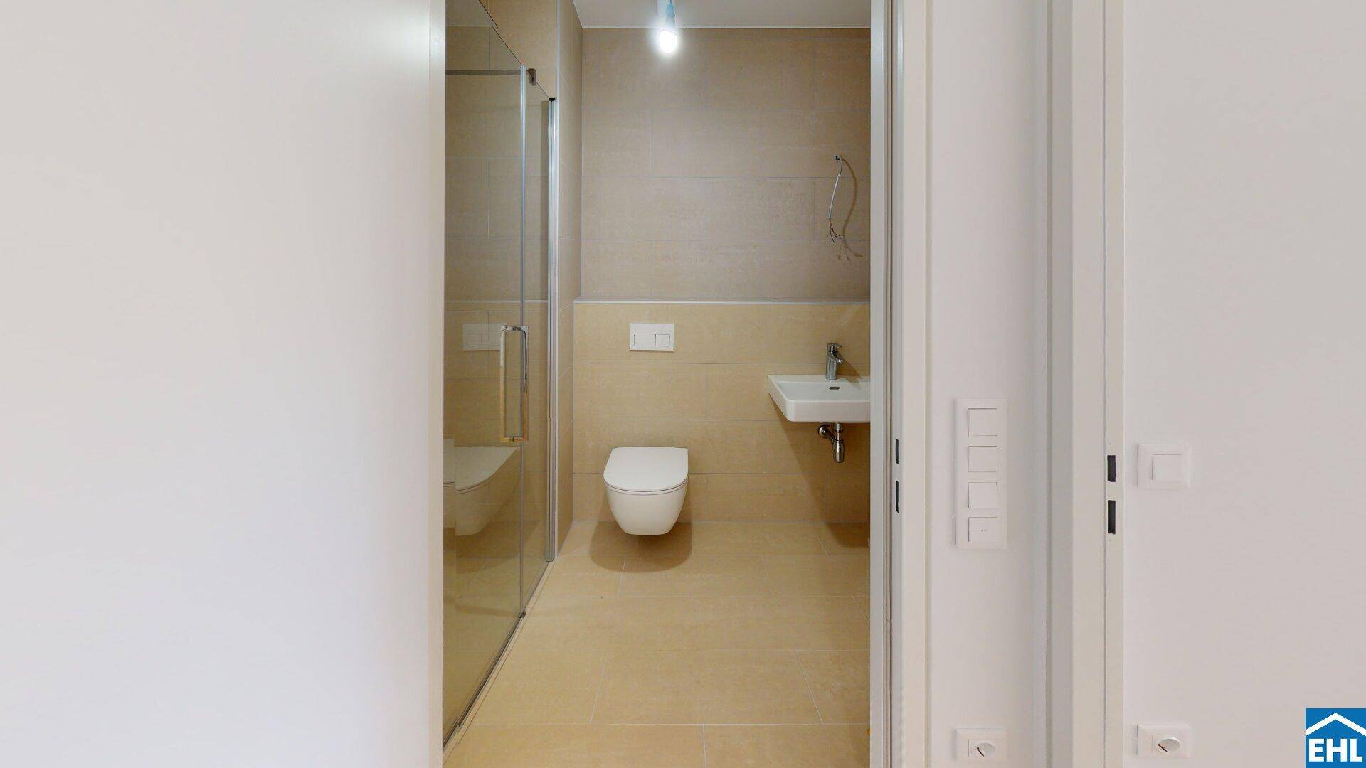 Funktionales Badezimmer mit wandhängendem WC, Duschkabine und einem kleinen Waschbecken.