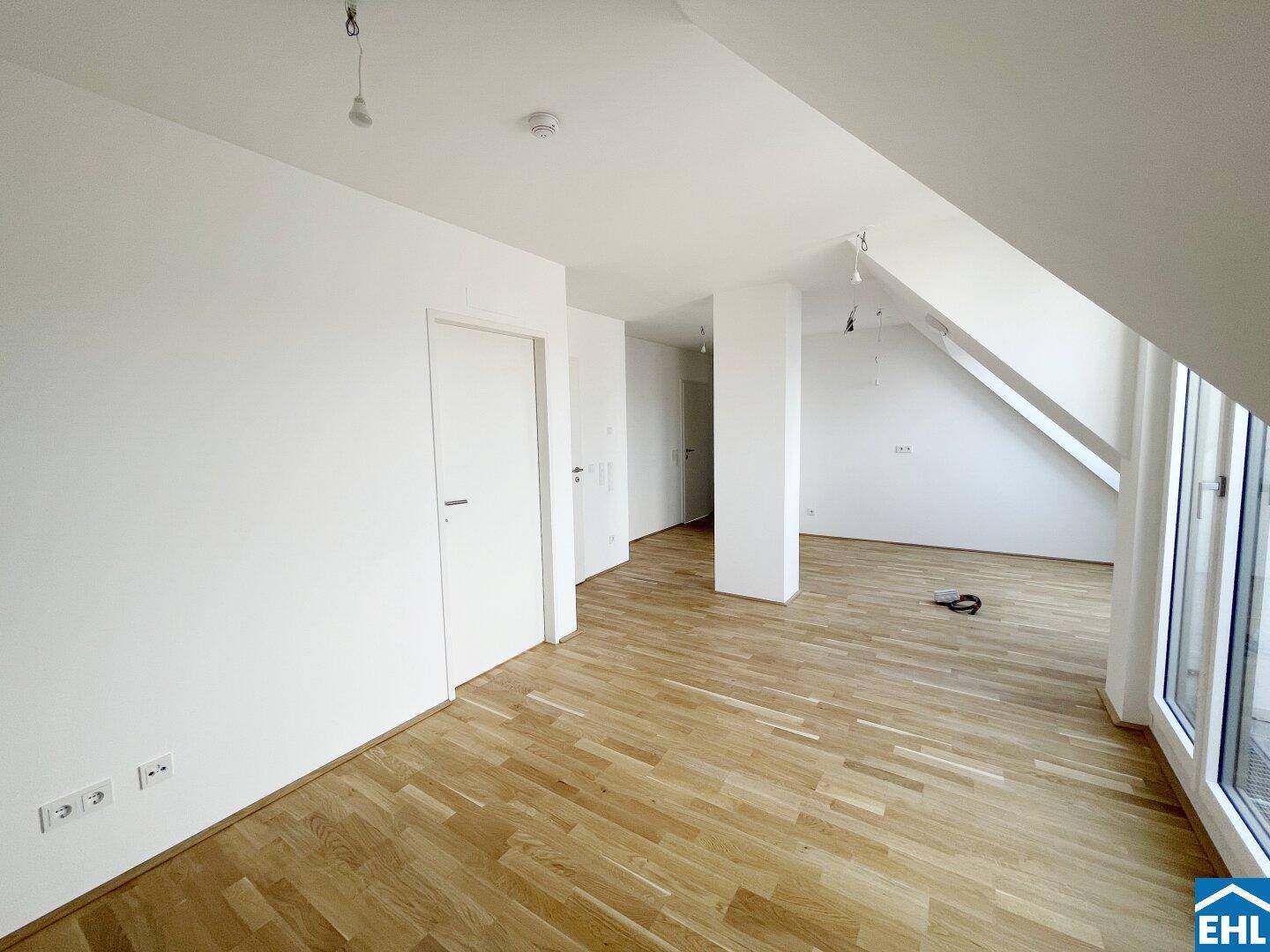 Leerer Dachgeschossraum mit Holzboden, weißen Wänden und Zugang zu einem Flur.