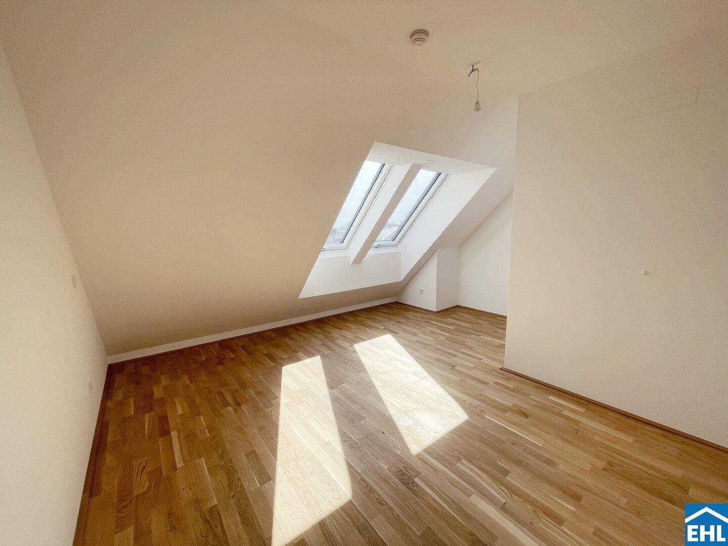 Heller Dachgeschossraum mit Holzboden und zwei schrägen Dachfenstern, die viel Sonnenlicht hereinlassen.