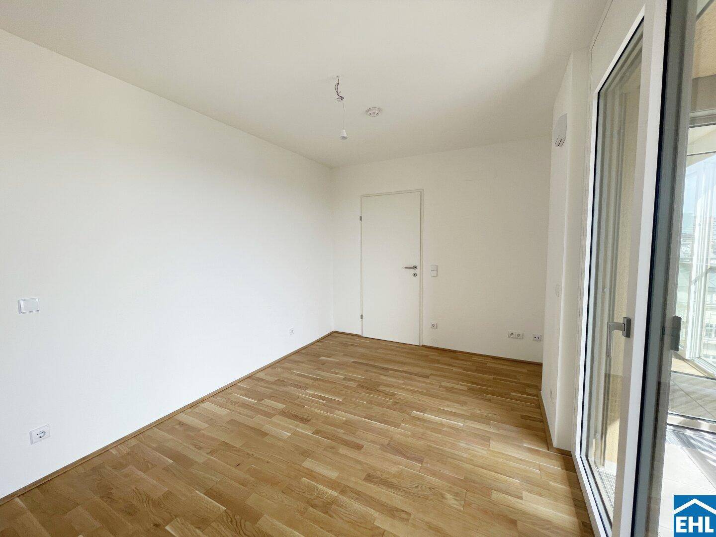 Unmöbliertes Zimmer mit Holzboden und großem Fenster, das viel Tageslicht hereinlässt.